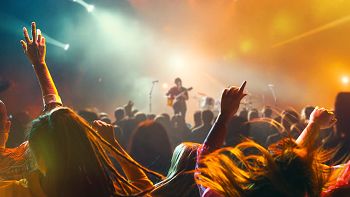 Multidão num concerto ao vivo, com braços erguidos em primeiro plano, palco ao fundo com artista em silhueta e luzes coloridas a iluminar o espaço.